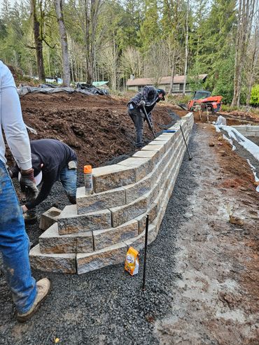Workers building a stone retaining wall outdoors in a wooded area.