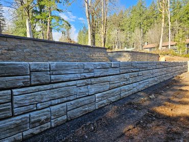 A newly built stone retaining wall in a wooded area under a clear sky.