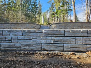 Stone retaining walls with a small stairway in a wooded area under a clear blue sky.