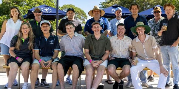 Group of people smiling and posing outdoors under a shaded area.