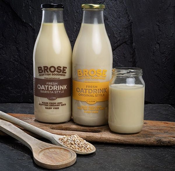 Two glass bottles of "Brose" oat milk, original and barista. Wooden spoons with oats lying in front. 