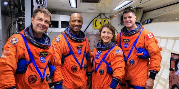 Four astronauts in orange NASA space suits posing inside a spacecraft.