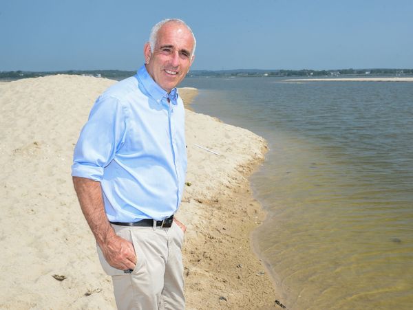 Man in light blue shirt standing on sandy shore by calm water under clear sky.