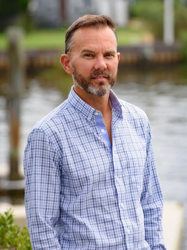Man in a blue checkered shirt standing near a waterfront.