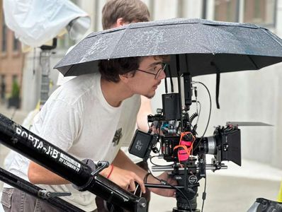 A person adjusting a professional camera under an umbrella on a rainy day.