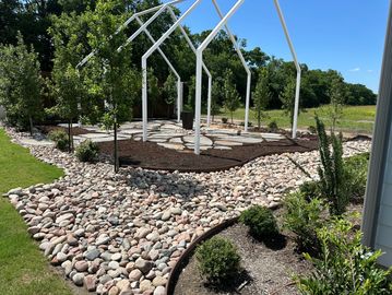 Landscaped garden with stone pathway and white metal frame structure under clear blue sky.