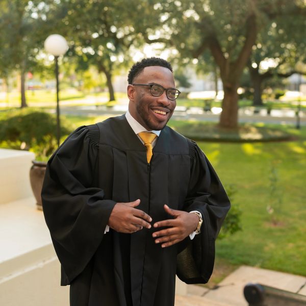 Smiling graduate in black gown and yellow tie outdoors on steps.