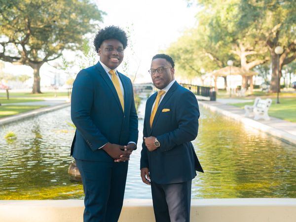 Two men in navy suits with yellow ties stand by a pond in a park.