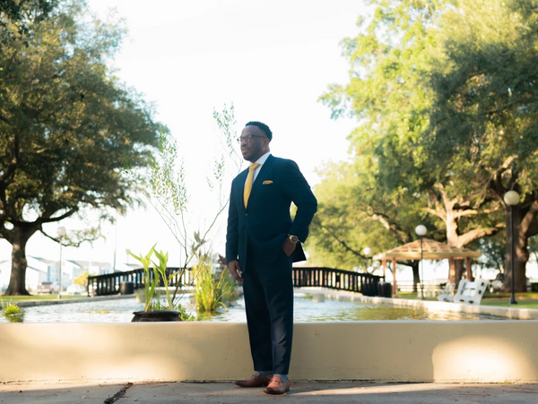 Man in a navy blue suit with a yellow tie standing by a fountain in a park.