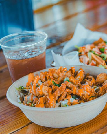 Crispy fried chicken bowl drizzled with spicy orange sauce and a cold drink on a wooden table.
