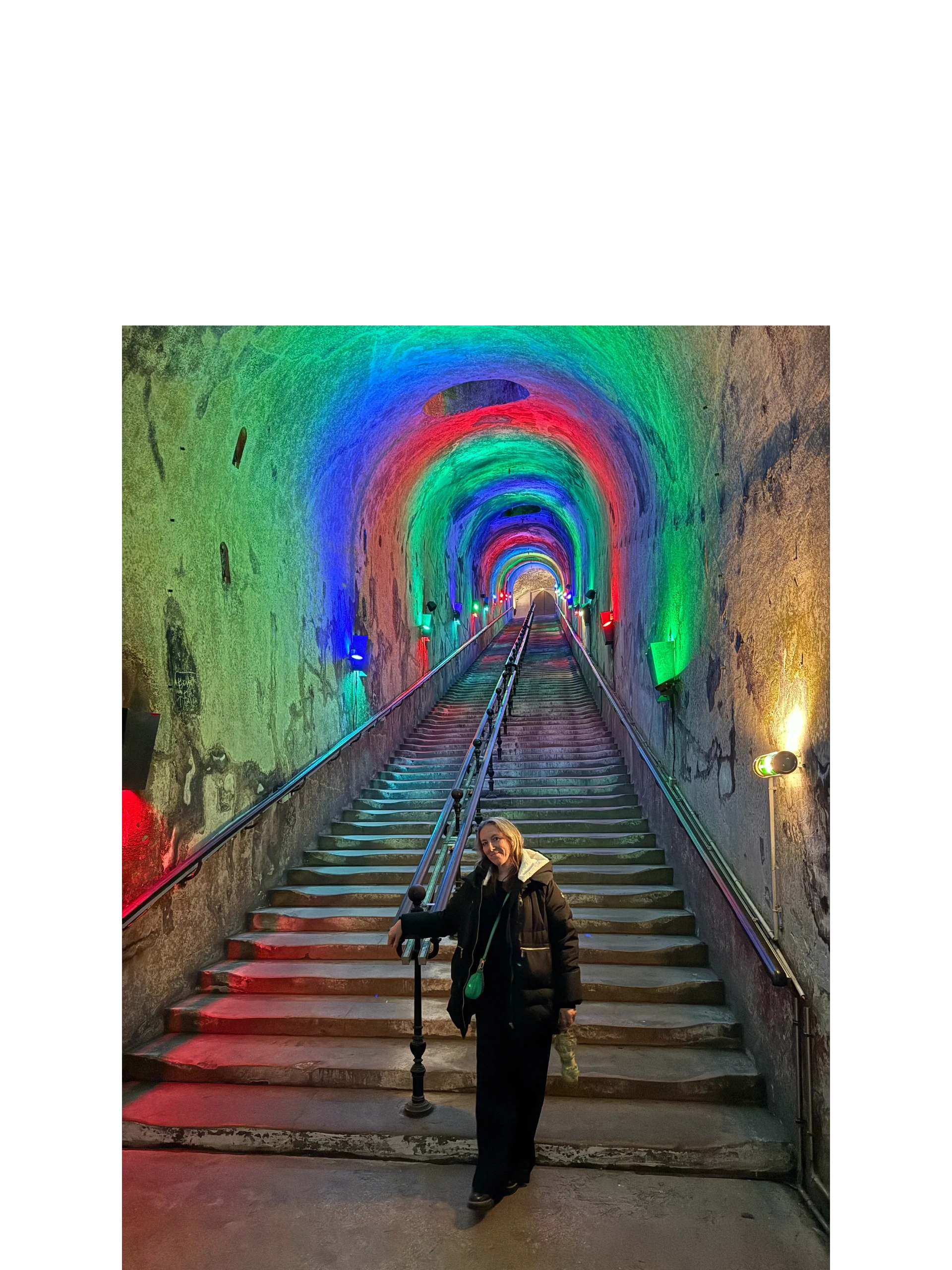 Woman standing at the base of a colorful, illuminated tunnel staircase.