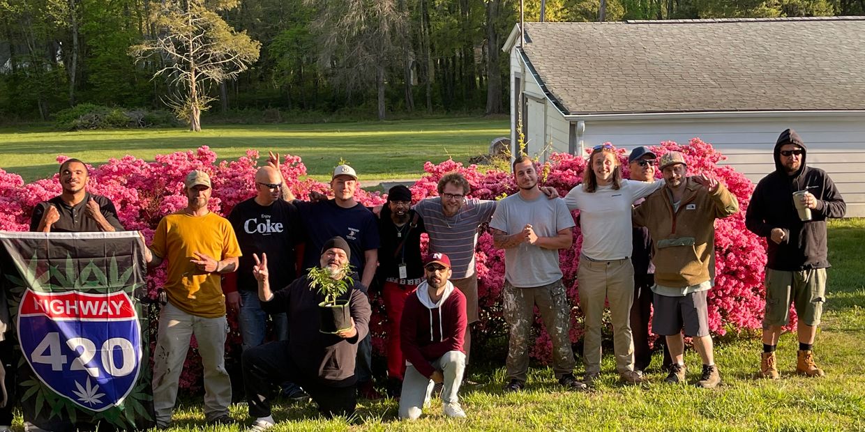 Group of friends posing outdoors with a 'Highway 420' flag and a potted plant, surrounded by pink flowers.
