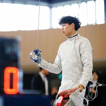 Young fencer in white gear holding foil, ready to compete indoors.