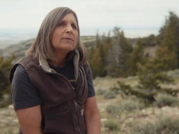 A woman in outdoor clothing sits in a natural, wooded area.