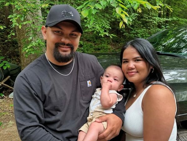 A smiling couple holding their baby outdoors with greenery in the background.
