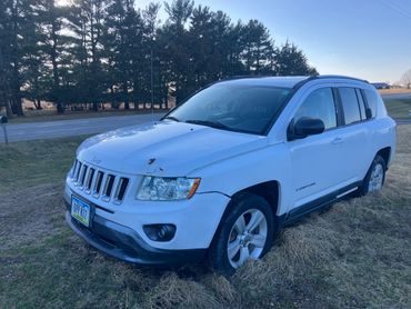 White Jeep Compass parked on grass near a road with some rust on the hood.