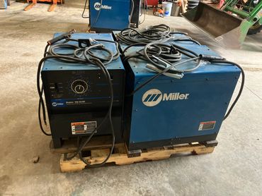 Two blue Miller welding machines on a wooden pallet in a workshop.
