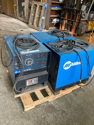 Two blue Miller welding machines on a wooden pallet in a workshop.