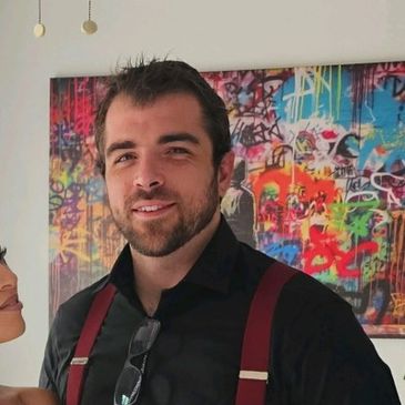 Man with dark hair and beard wearing suspenders stands in front of colorful graffiti art.