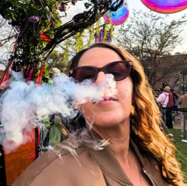 Person wearing sunglasses blowing smoke outdoors at a colorful event.