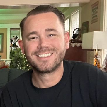 Smiling man with short hair and a beard in a cozy indoor setting.