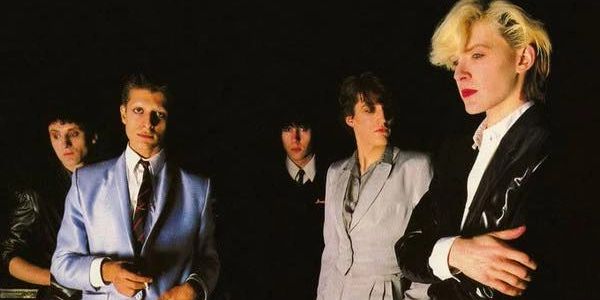 A 1980s band posing with stylish, vintage outfits against a dark background.