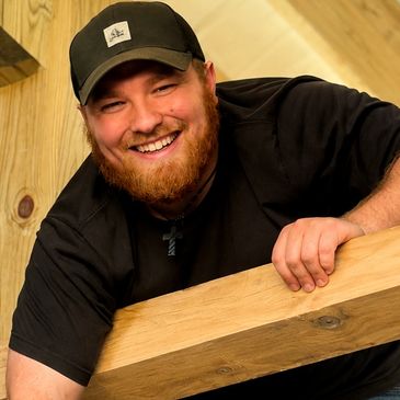 Smiling man with a beard and cap holding a wooden plank indoors.