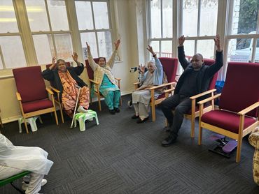 Four elderly people sitting on chairs with arms raised indoors.