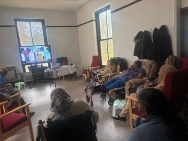 Elderly people watching TV together in a cozy room with chairs and wheelchairs.