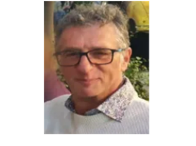 Man with glasses smiling outdoors wearing a white sweater.
