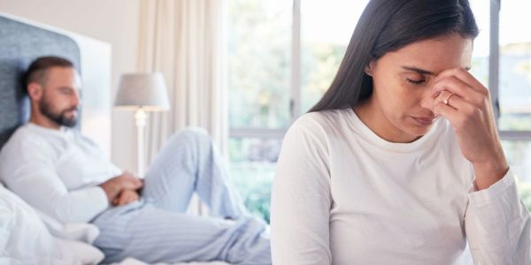 Sad woman covering eyes, man sitting on bed in background.