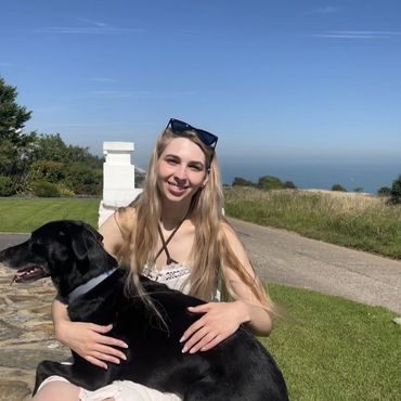 A young woman smiles while hugging her black dog outdoors on a sunny day.
