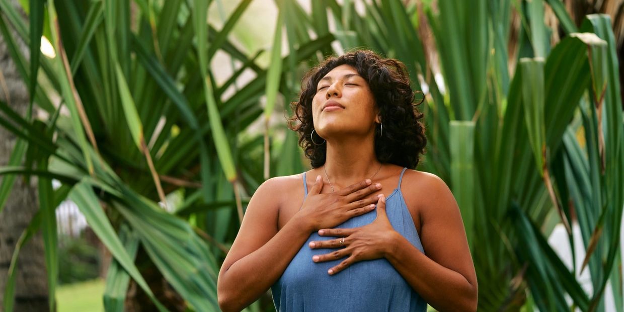 Woman in blue dress with eyes closed, hands on chest, surrounded by green plants.