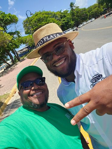 Two men smiling outdoors on a sunny day, one wearing a straw hat.