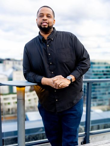 Man in black shirt leaning on a rooftop railing with cityscape background.