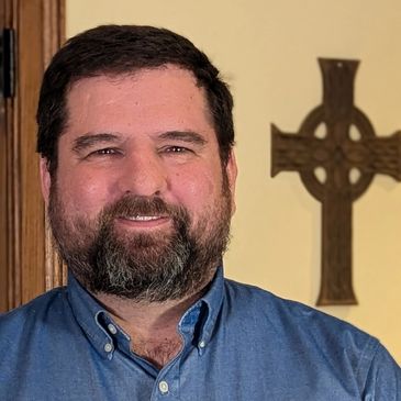 Smiling man with beard in blue shirt standing indoors with a decorative cross on the wall.