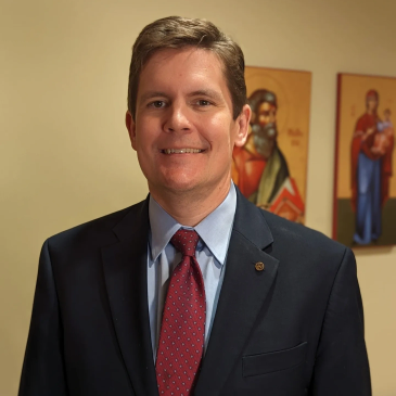 Smiling man in suit and red tie with religious paintings in background.