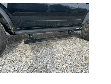 Close-up of a black truck with extended power step side rails on gravel.