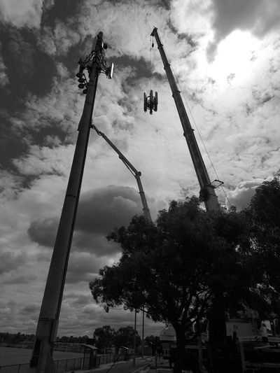 Cranes lifting equipment onto a tall pole under a cloudy sky.