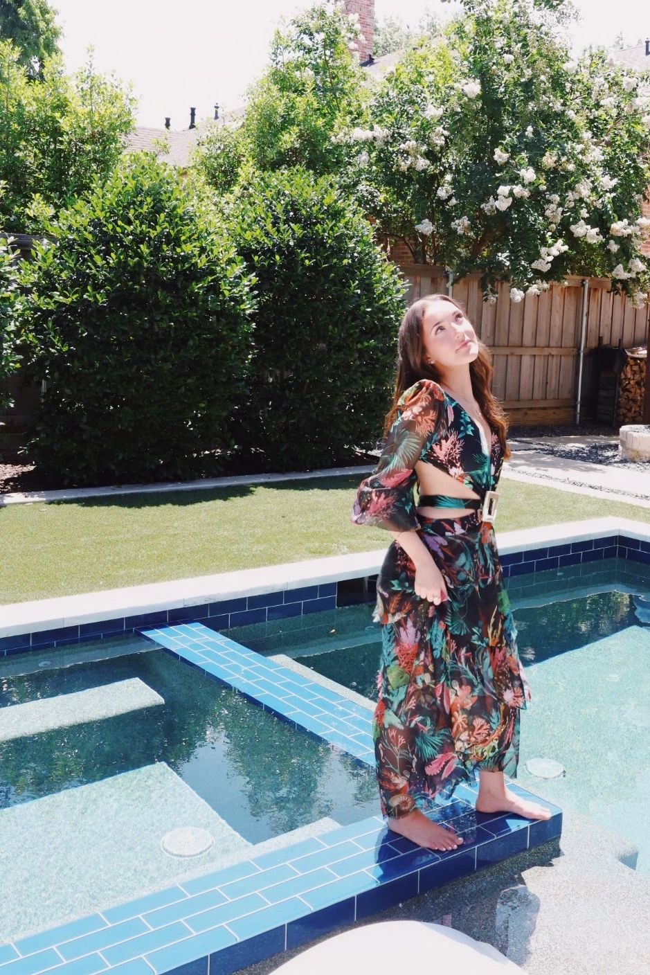 Woman in floral dress standing on edge of a pool in a backyard.