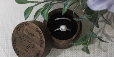 A wooden ring box with two sparkling rings inside, surrounded by greenery.