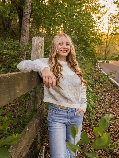 Young woman in a white sweater leaning on a wooden fence outdoors.