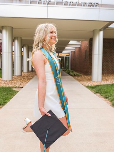 Young woman in white dress proudly holding graduation cap outdoors.