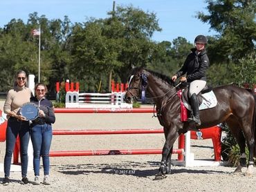 Equestrian rider on horse with two women holding a trophy beside a jump.
