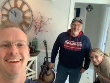 Three people smiling inside a cozy room with a guitar and wall clock.