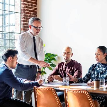 A group of people at a meeting table with an older man standing at the head gesturing to something.