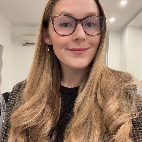 A young woman with long blonde hair and tortoiseshell glasses smiling indoors.
