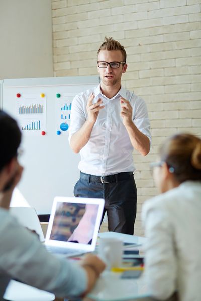 A man gestures animatedly while presenting in a modern office, with charts displayed on a board