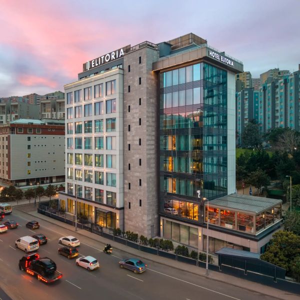Modern Elitoria Hotel with glass facade at sunset beside busy road.