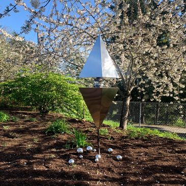 Shiny geometric garden sculpture with cherry blossom trees in the background.